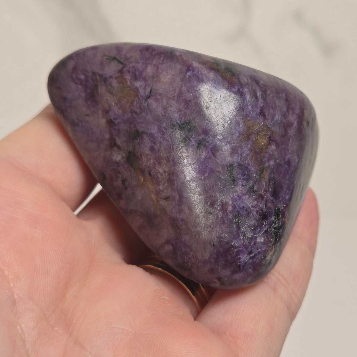 A polished purple tumbled stone, likely amethyst, held in a person's hand against a light marble-patterned background.