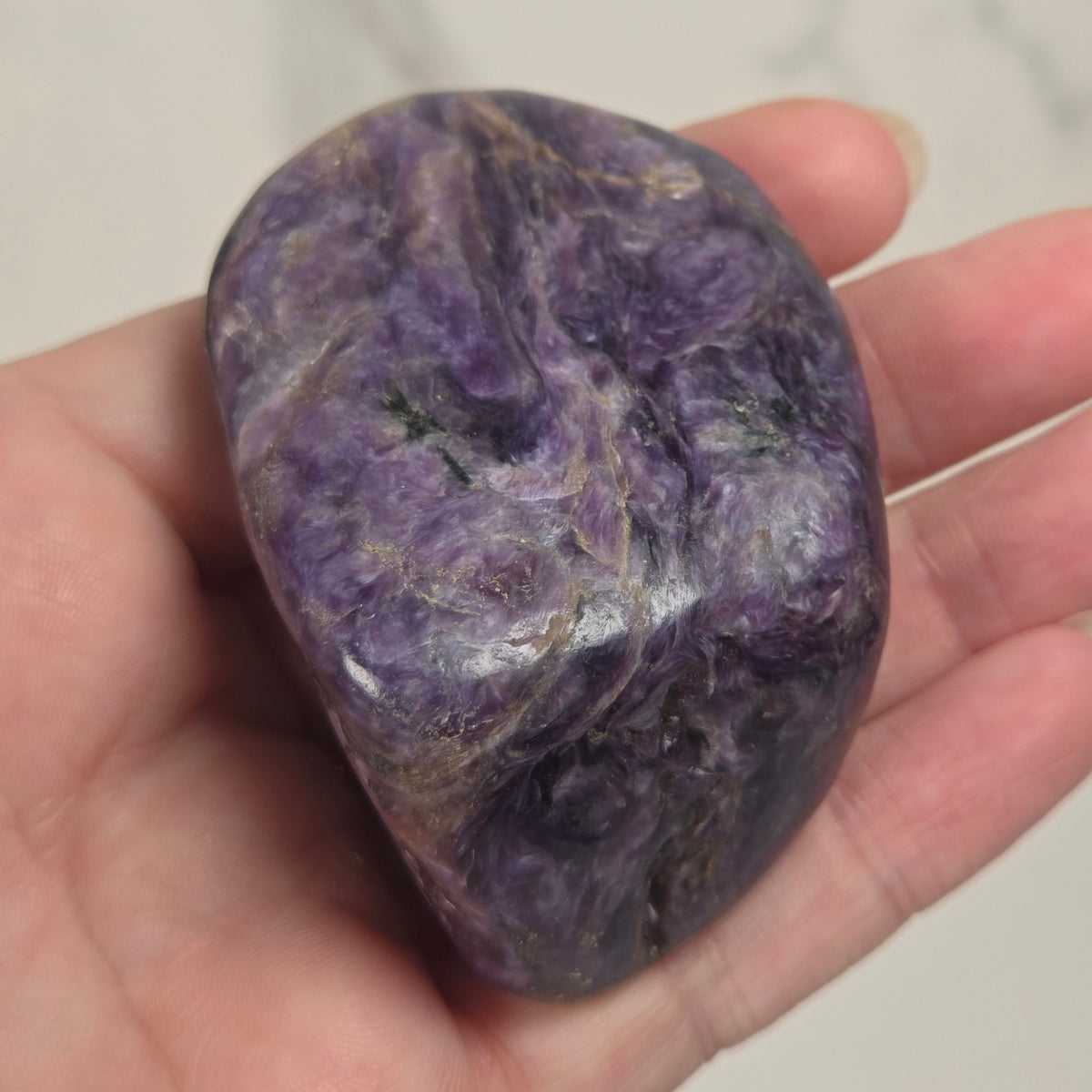 A polished purple amethyst stone held in a person's hand against a light marble-patterned background.