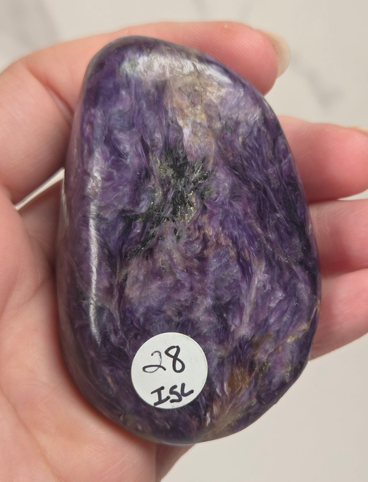 A hand holding a polished purple tumbled stone with a small white sticker reading '28 ISK' on a light neutral background.