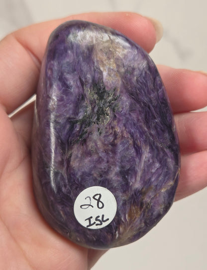 A hand holding a polished purple tumbled stone with a small white sticker reading '28 ISK' on a light neutral background.