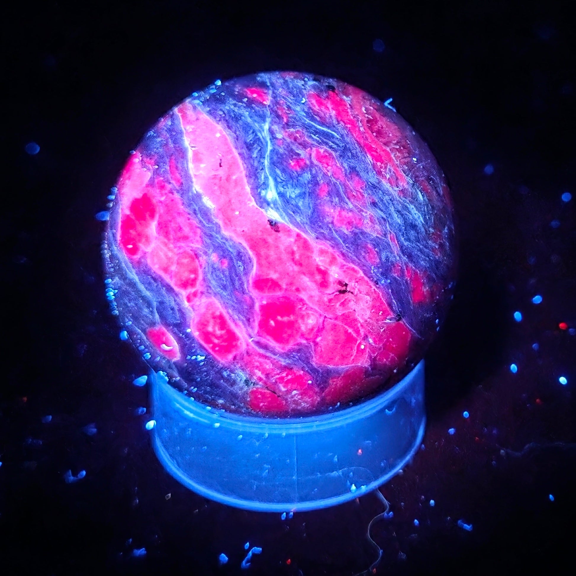 A spherical  orb with a vibrant pink and blue marbled pattern, displayed on a blue circular base against a black background under UV light.