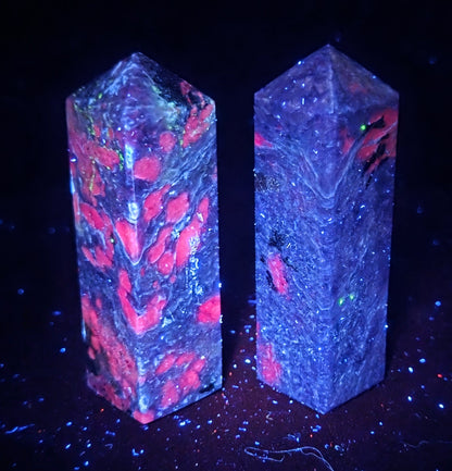 Two rectangular crystal towers with pointed tops, featuring a blacklight-reactive design with pink, red, and green inclusions under UV light, set against a dark black background.