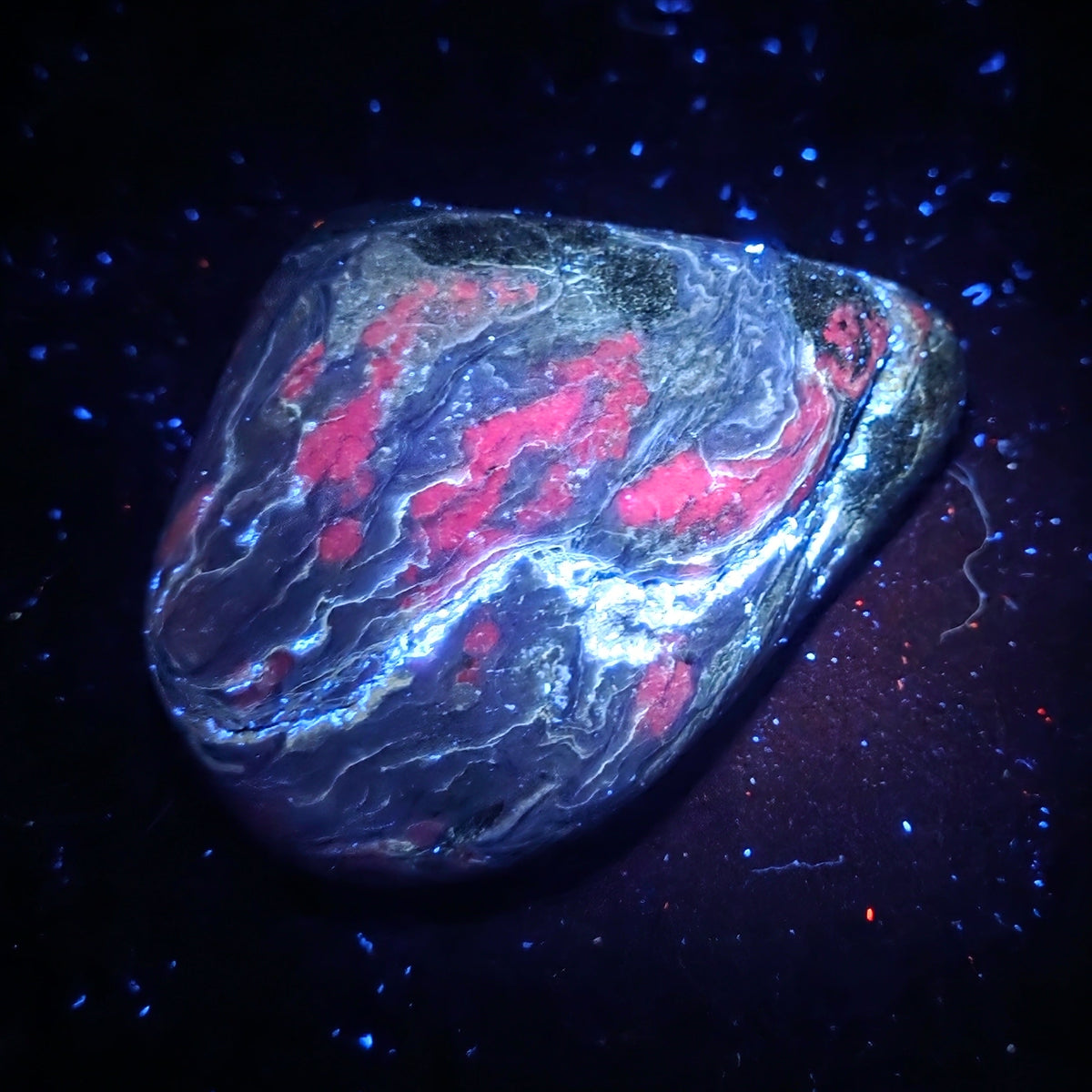 A polished decorative stone with a marbled pattern of black, blue, white, and bright pink, photographed on a dark background with blue sparkles.