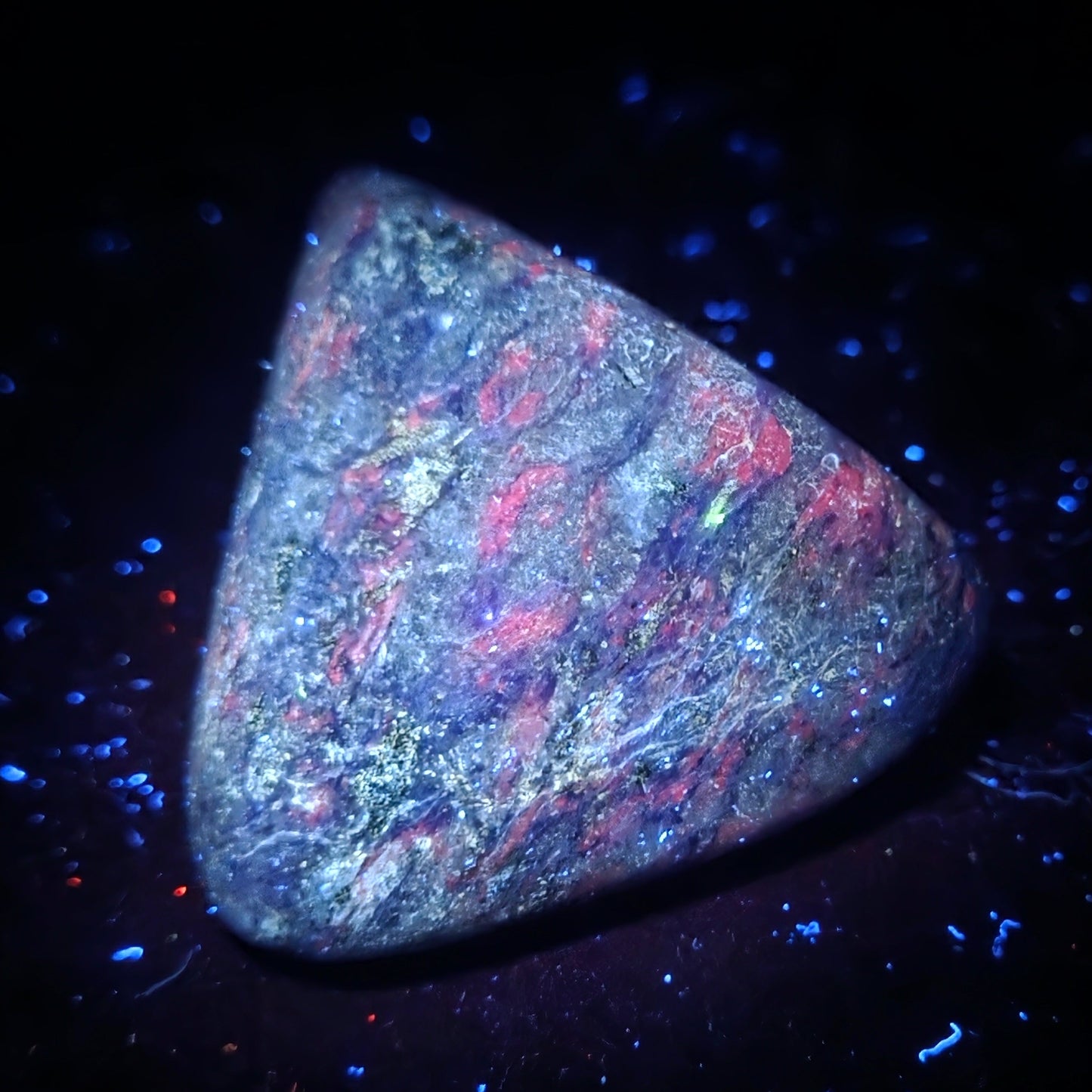 A triangular polished gemstone or opalite specimen with a mottled play-of-color surface, showing blue, pink, purple, and gray tones, photographed on a black background with scattered blue and red fluorescent specks.