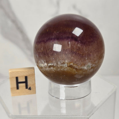 A polished purple amethyst crystal sphere on a clear acrylic stand, next to a Scrabble tile showing the letter H, set against a light gray marble background.