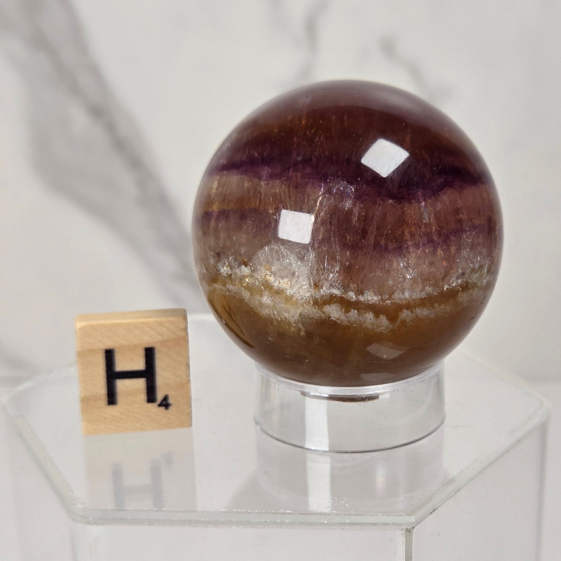 A polished brown and purple crystal sphere with natural banding, displayed on a clear acrylic stand next to a wooden Scrabble tile showing the letter 'H'.