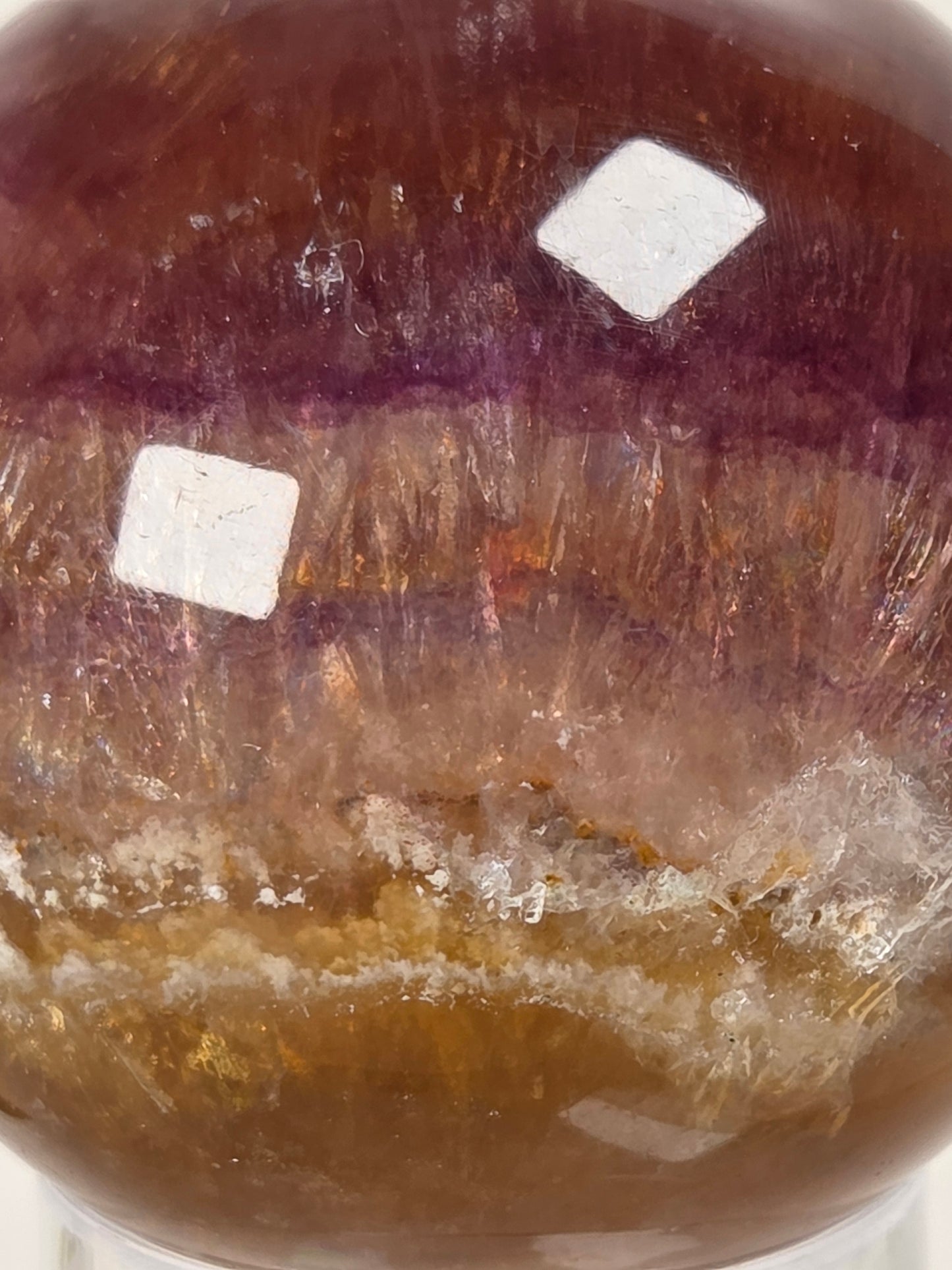 A close-up of a polished round crystal sphere showing bands of amber, purple, and translucent golden tones, with two white square stickers on the surface.