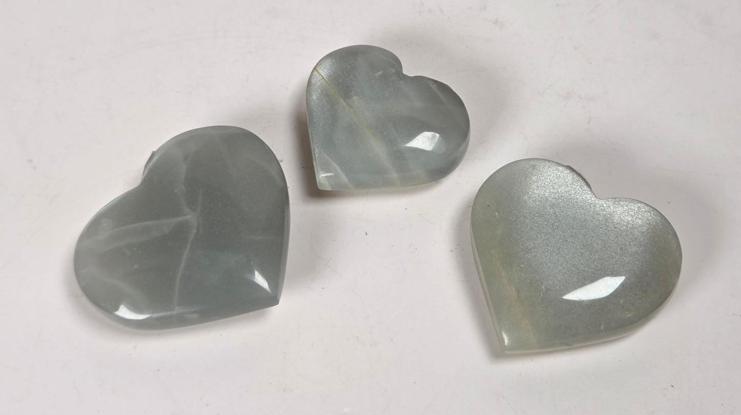 Three heart-shaped translucent cabochon gemstones in a pale gray-green hue, arranged on a plain off-white surface.