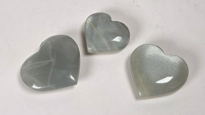 Three heart-shaped translucent cabochon gemstones in a pale gray-green hue, arranged on a plain off-white surface.