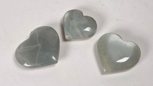 Three heart-shaped translucent cabochon gemstones in a pale gray-green hue, arranged on a plain off-white surface.