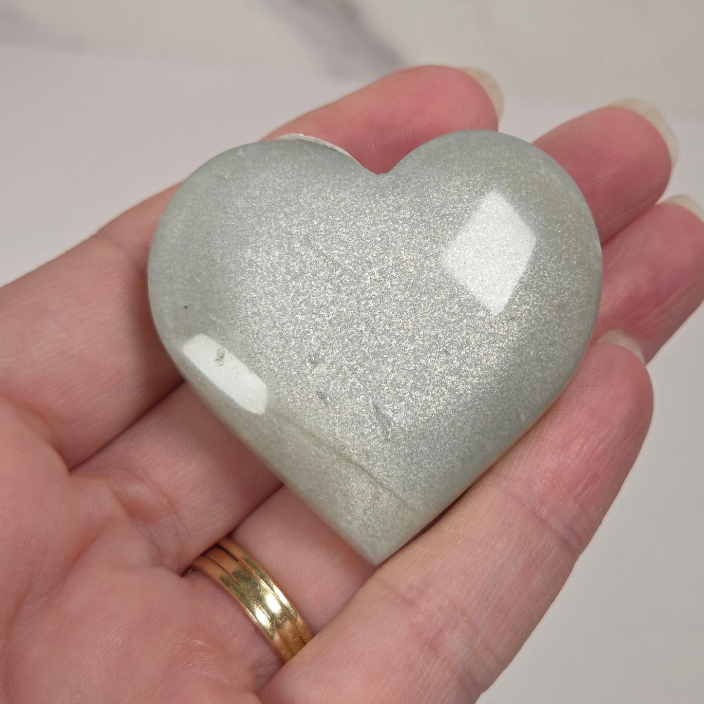 A polished, heart-shaped stone with a light gray and white glittery finish, held in a person's hand against a light marble-patterned background.