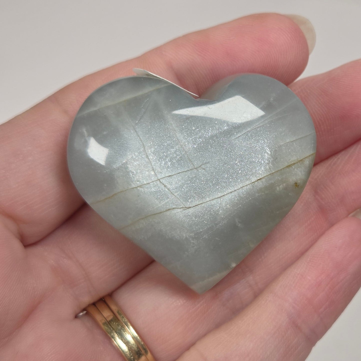 A heart-shaped polished stone held in a person's hand. The stone has a smooth, glossy finish with natural banding and is a pale gray-green color.