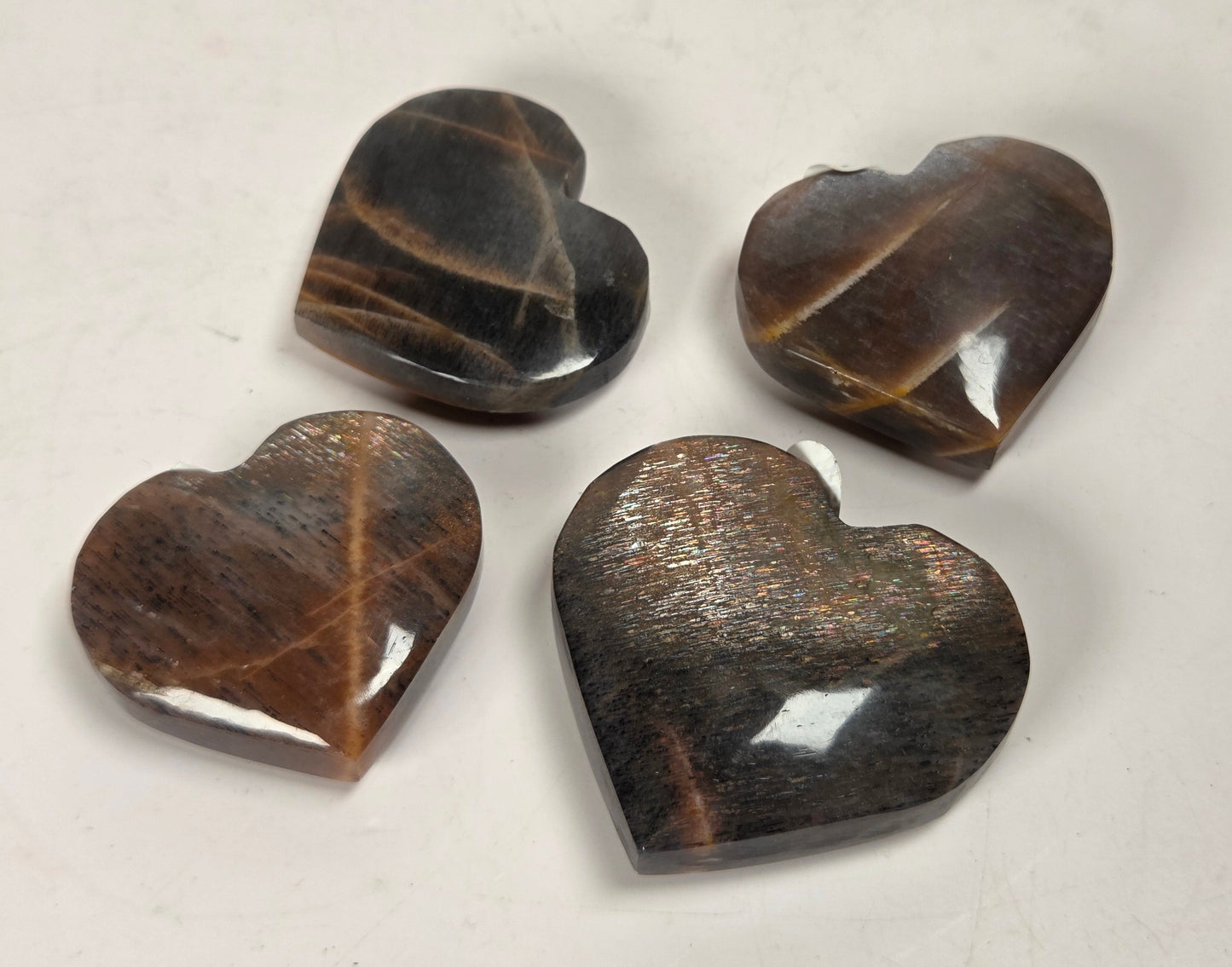 Four polished heart-shaped gemstone pendants in brown and black tiger's eye stone, arranged on a light off-white background.