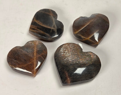 Four polished heart-shaped gemstone pendants in brown and black tiger's eye stone, arranged on a light off-white background.