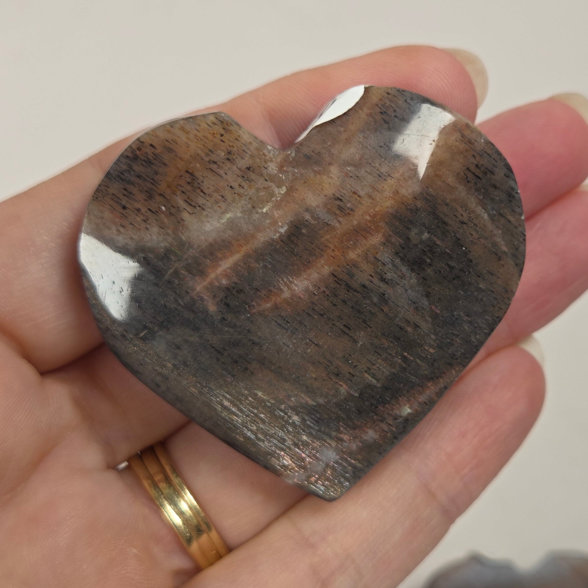 A hand holding a polished heart-shaped brown stone with natural banding, with another similar stone blurred in the background.