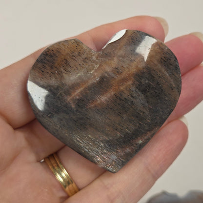 A hand holding a polished heart-shaped brown stone with natural banding, with another similar stone blurred in the background.
