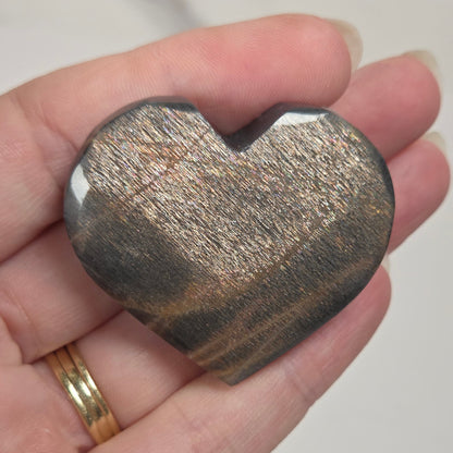 A heart-shaped polished stone with a metallic gold and dark gray banded pattern, held in a person's hand.
