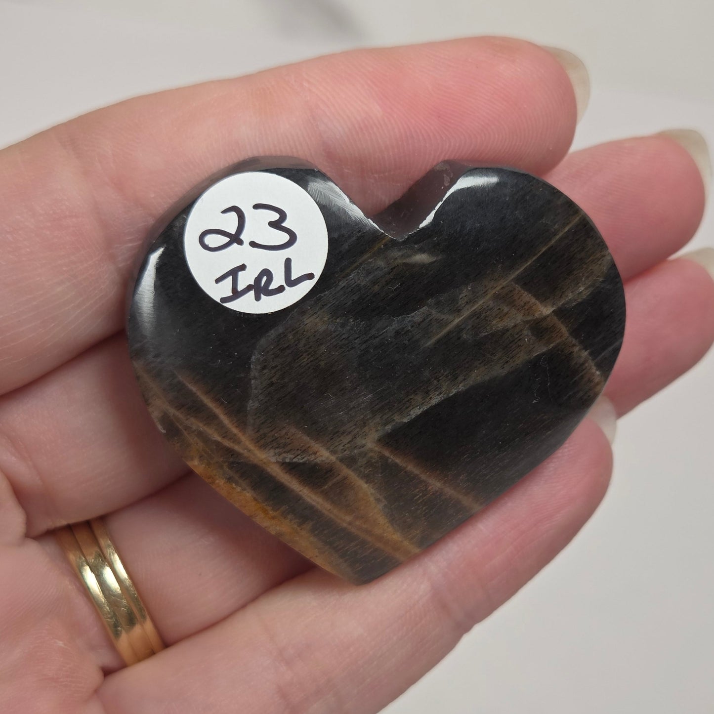 A heart-shaped polished black stone with brown veining, held in a person's hand. A small white sticker on the stone reads '23 Irl'.