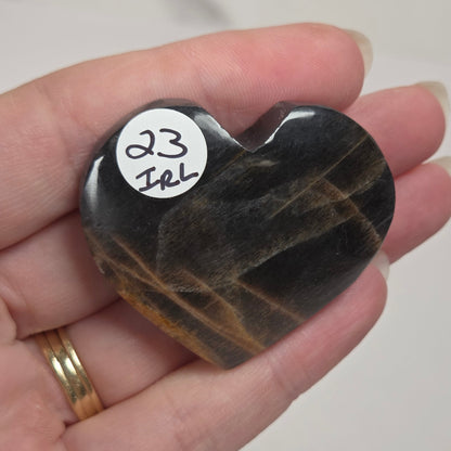 A heart-shaped polished black stone with brown veining, held in a person's hand. A small white sticker on the stone reads '23 Irl'.