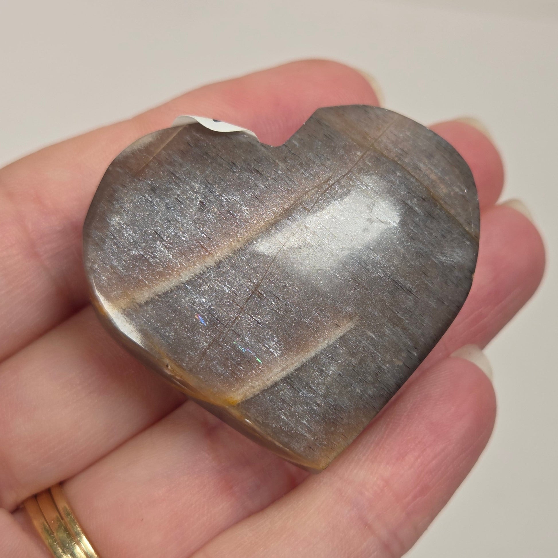 A heart-shaped polished stone with a metallic silver-gray finish and natural striations, held in a person's hand. The hand has a gold ring on one finger, and the background is a clean, neutral off-white surface.