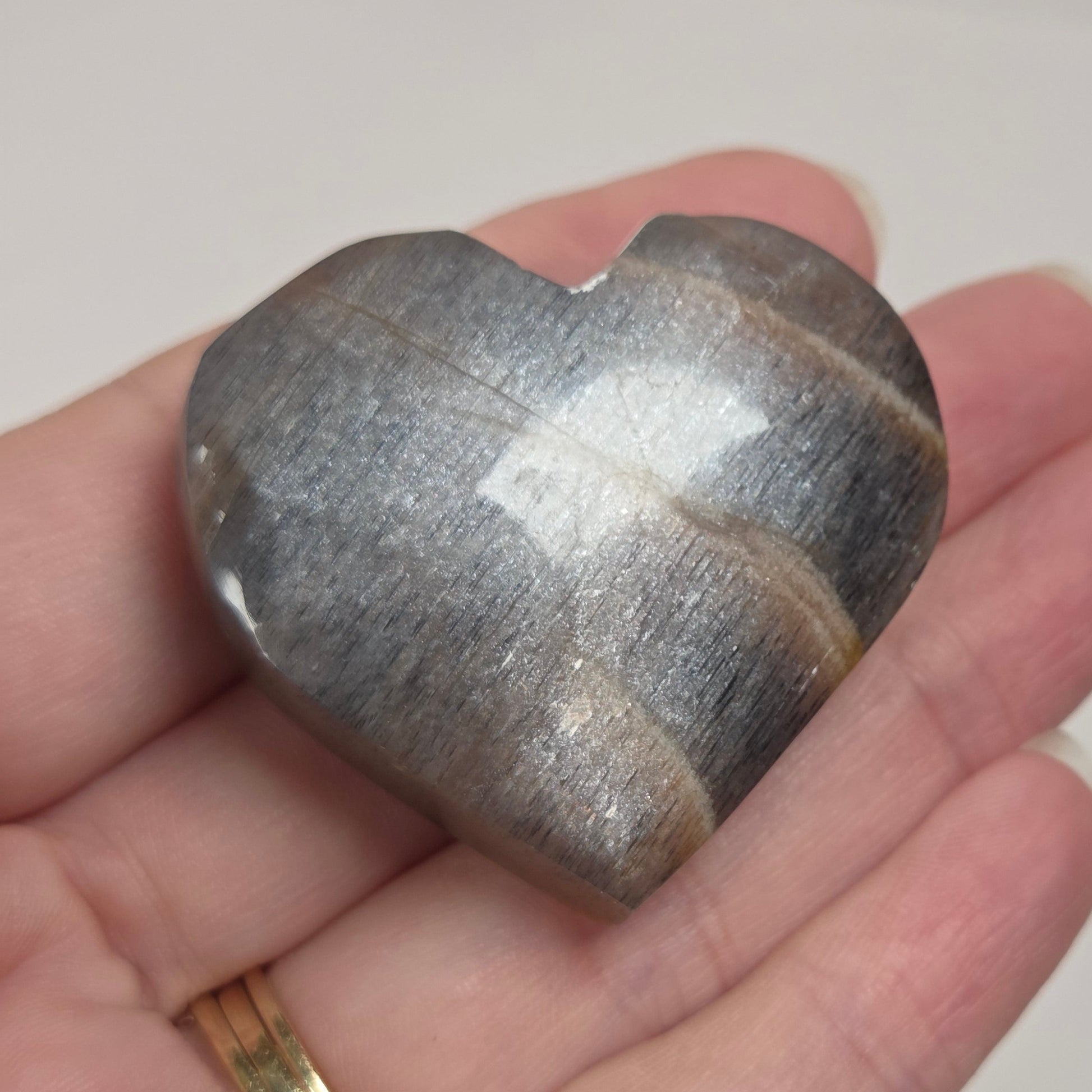 A polished heart-shaped stone with natural banding, held in a person's hand. The stone has a metallic silver-gray appearance with darker brownish-gray bands, and the hand is wearing a gold ring.