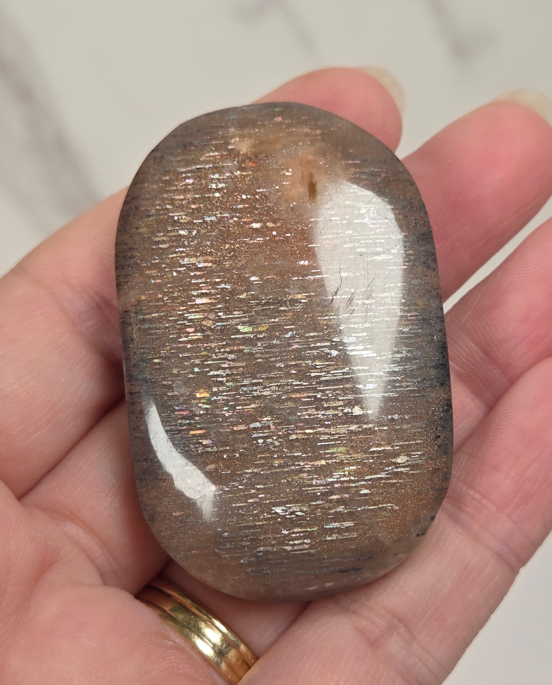 A polished brown tumbled stone with fine metallic gold inclusions, held in a person's hand against a light marble-patterned background.