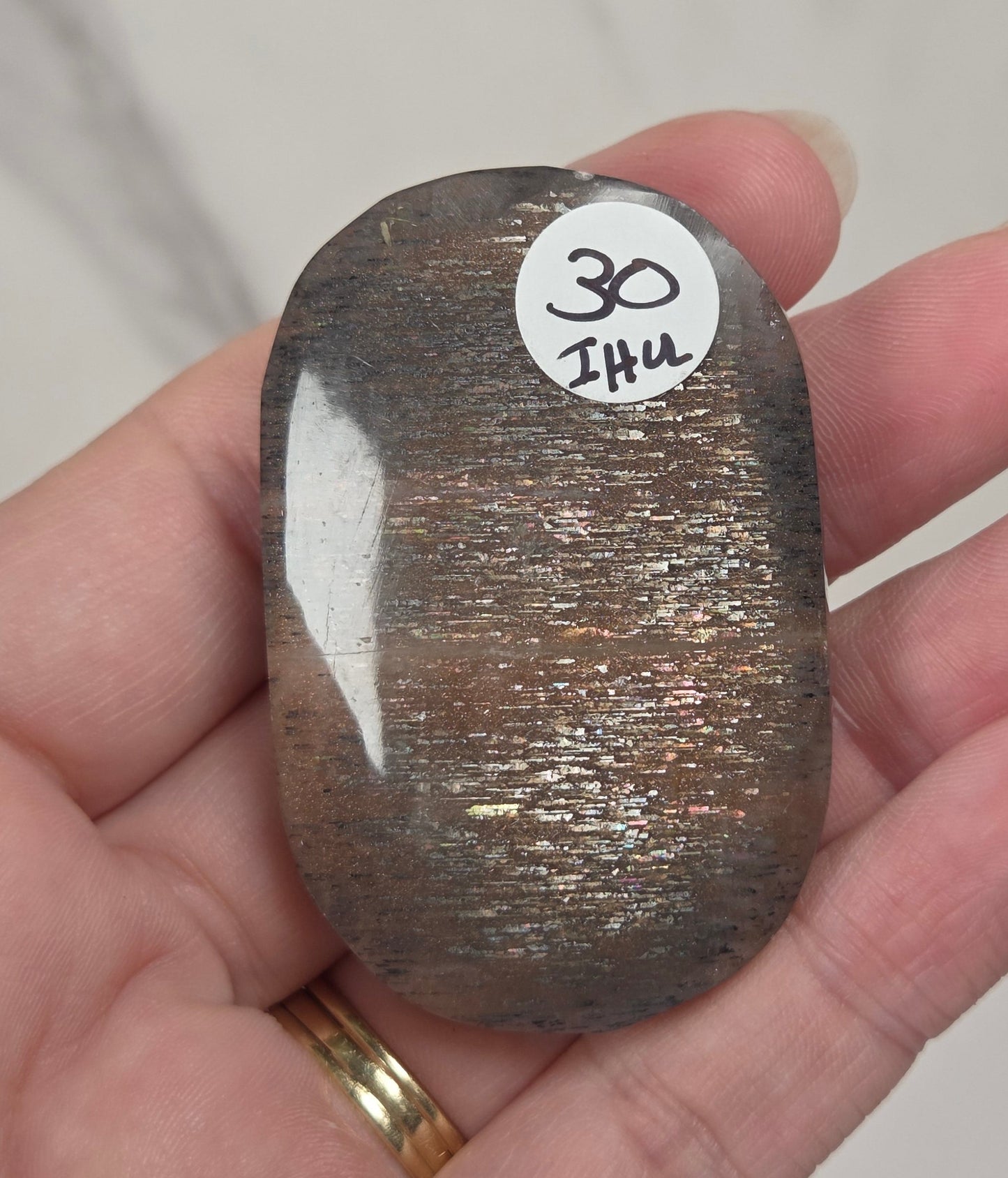 A hand holds a polished, smooth stone with a metallic bronze-brown sheen and fine glitter, featuring a small white sticker with the text '30 Zhu'.