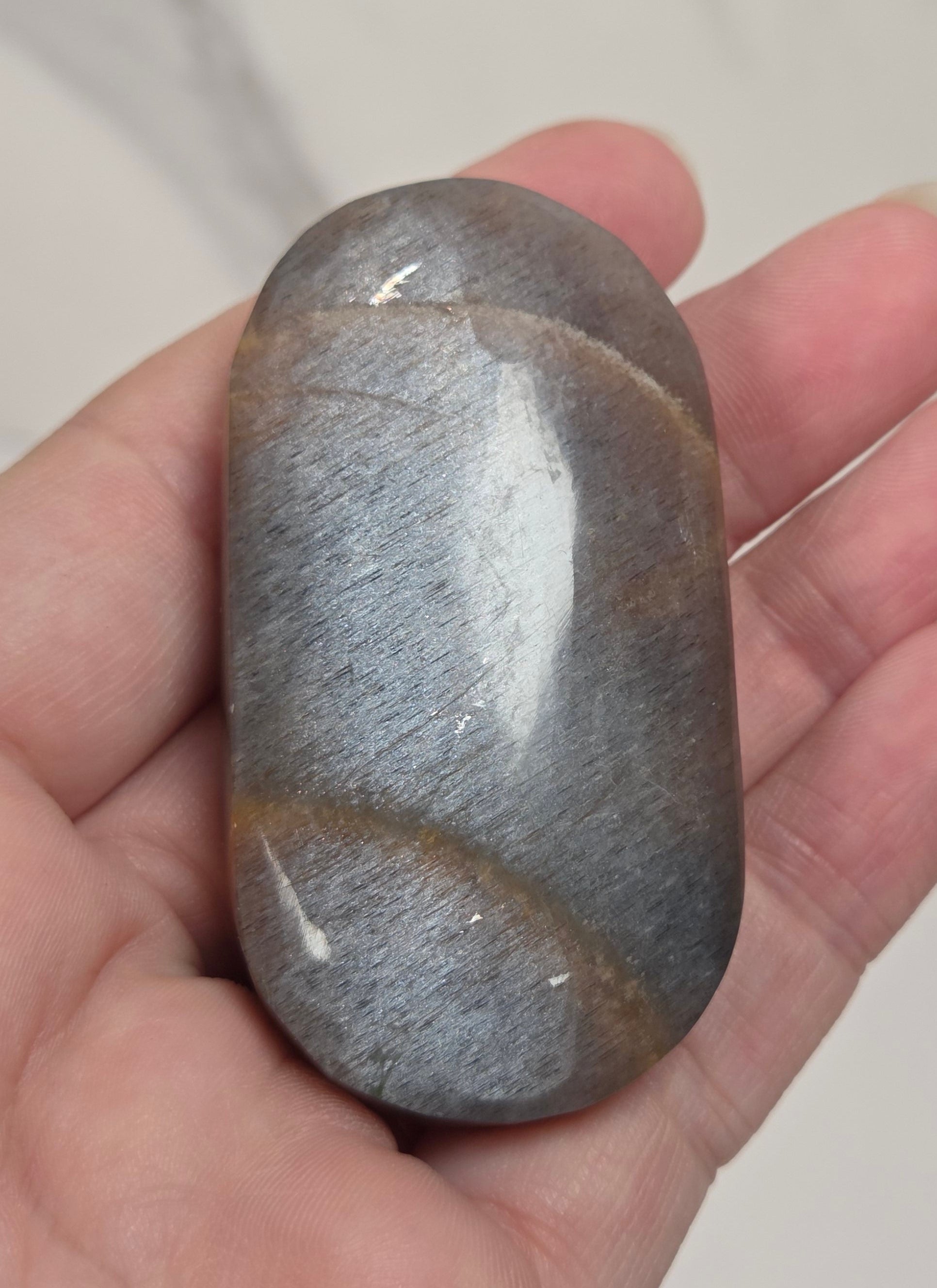 A hand holding a polished gray stone with subtle brown veining, set against a light marble-patterned background.