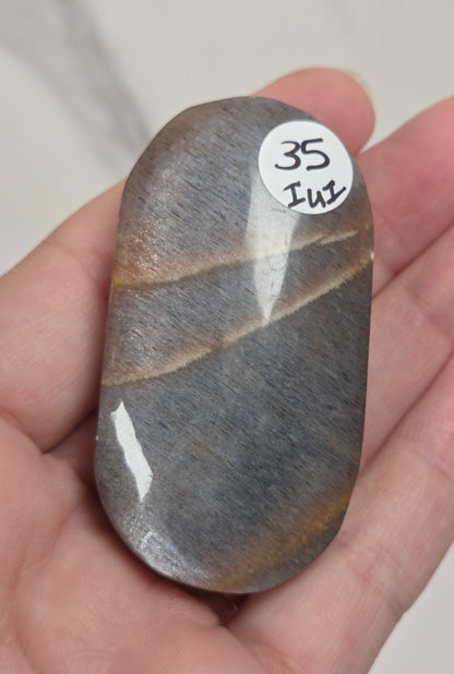 A hand holding a polished gray stone with natural brown veining and a small white sticker labeled '35 IVI' on its surface, set against a light marble-patterned background.