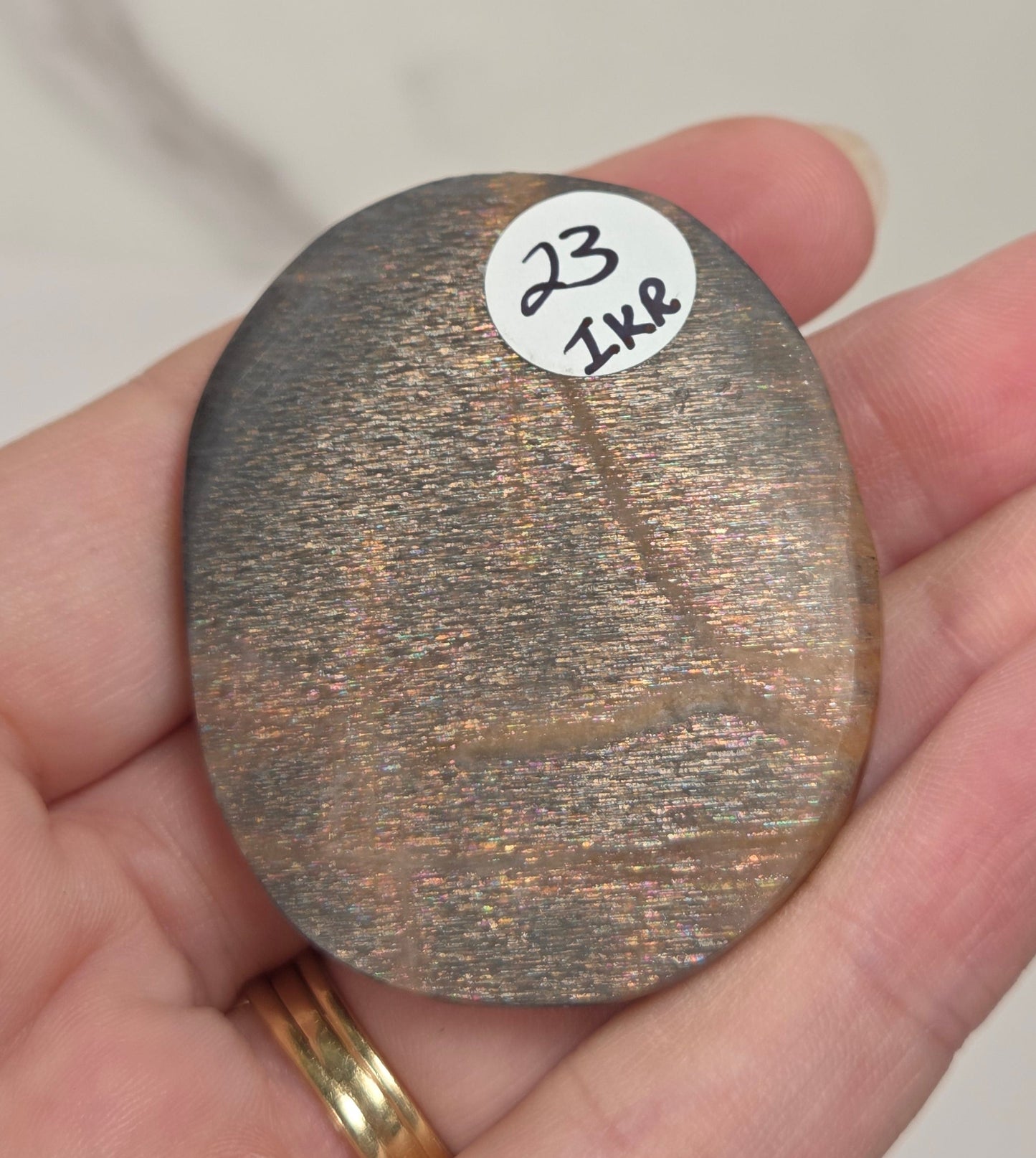 A hand holding a small, oval, metallic bronze-colored stone or chip with a white sticker labeled '23 1K8' on it, set against a light marble background.