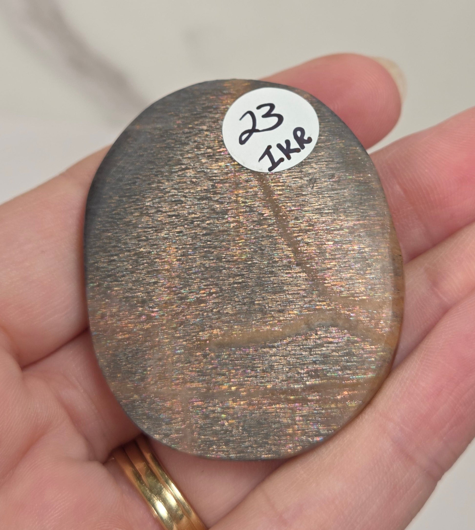 A hand holding a small, oval, metallic bronze-colored stone or chip with a white sticker labeled '23 1K8' on it, set against a light marble background.