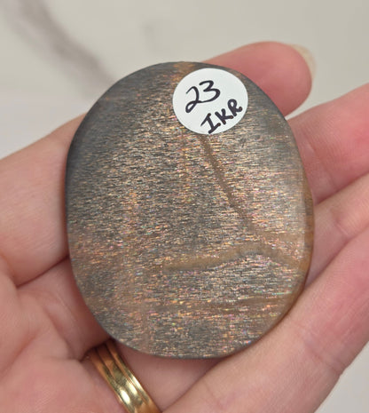 A hand holding a small, oval, metallic bronze-colored stone or chip with a white sticker labeled '23 1K8' on it, set against a light marble background.