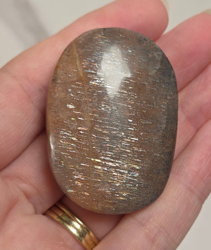 A polished brown stone with fine metallic-looking striations, held in a person's hand against a light marble-like background.