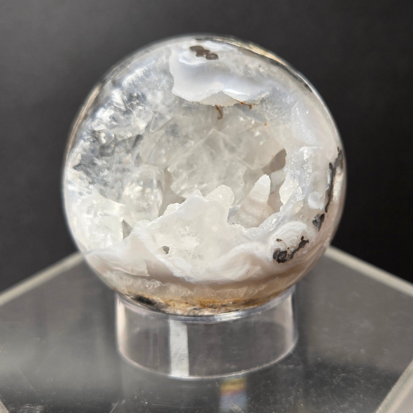 A clear crystal geode sphere with white agate formations and dark inclusions, displayed on a clear acrylic stand against a dark gray background.