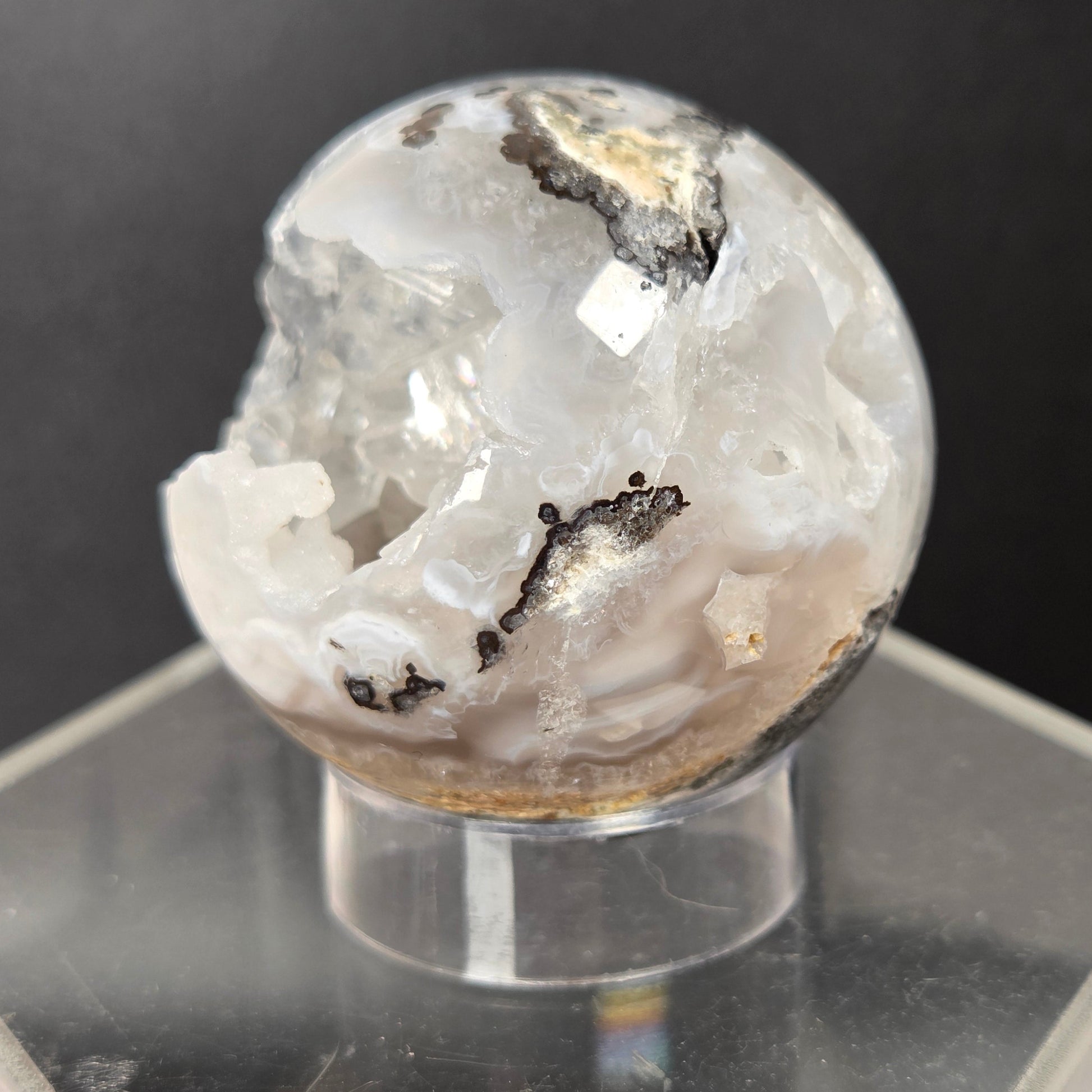 A spherical agate crystal geode specimen with white, clear, and gray crystalline patterns and dark inclusions, displayed on a clear acrylic hexagonal stand against a dark gray background.