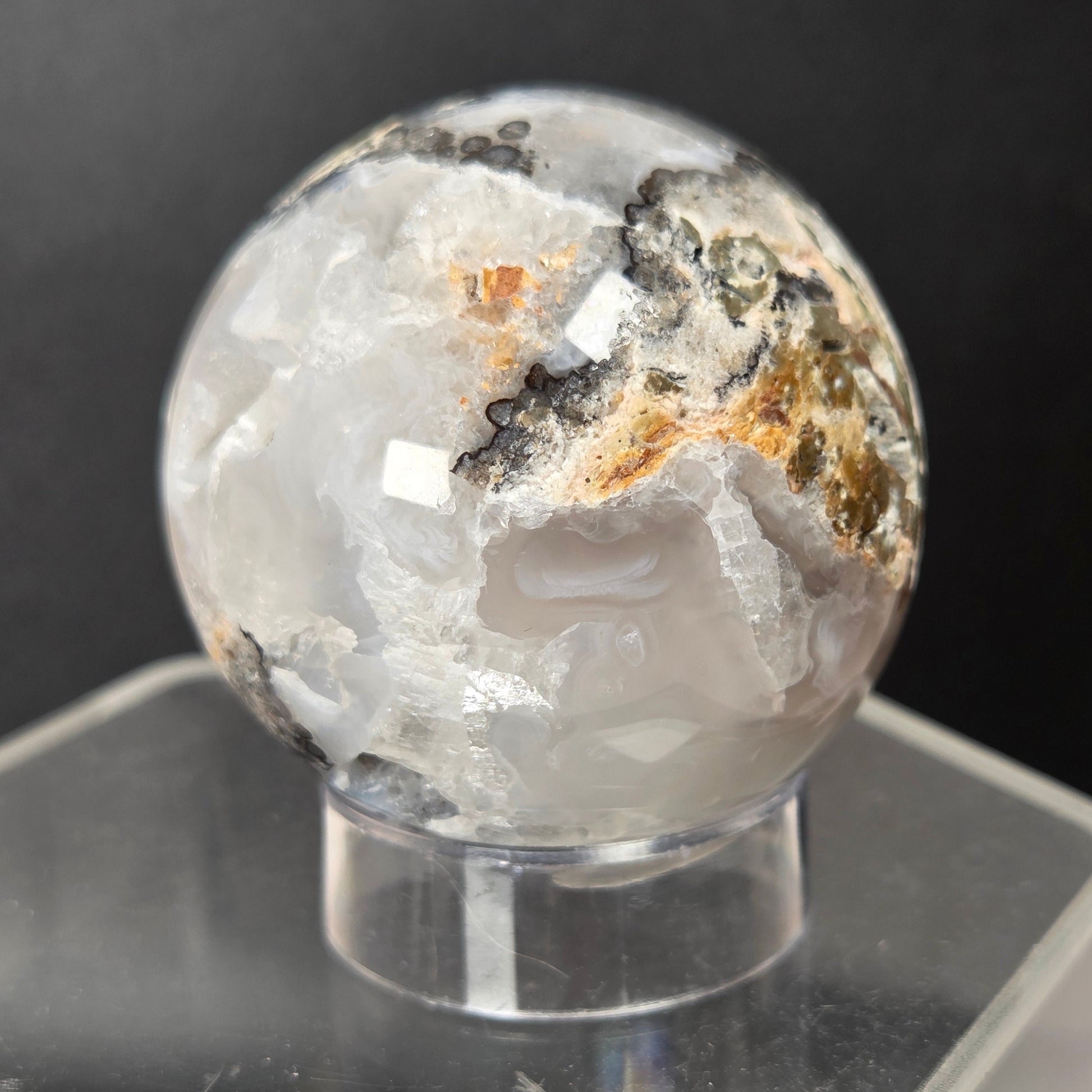 A polished crystal sphere with white, clear, and gray agate-like formations and small brown and black mineral inclusions, displayed on a clear acrylic stand against a dark gray background.
