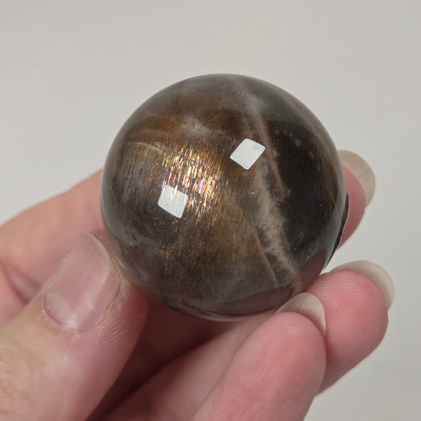 A hand holding a polished round brown stone with a metallic gold sheen and white banding, set against a light gray background.