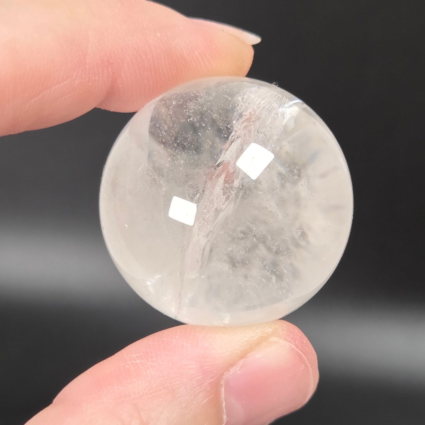 A hand holding a small, polished, transparent white stone sphere against a dark, out-of-focus background.
