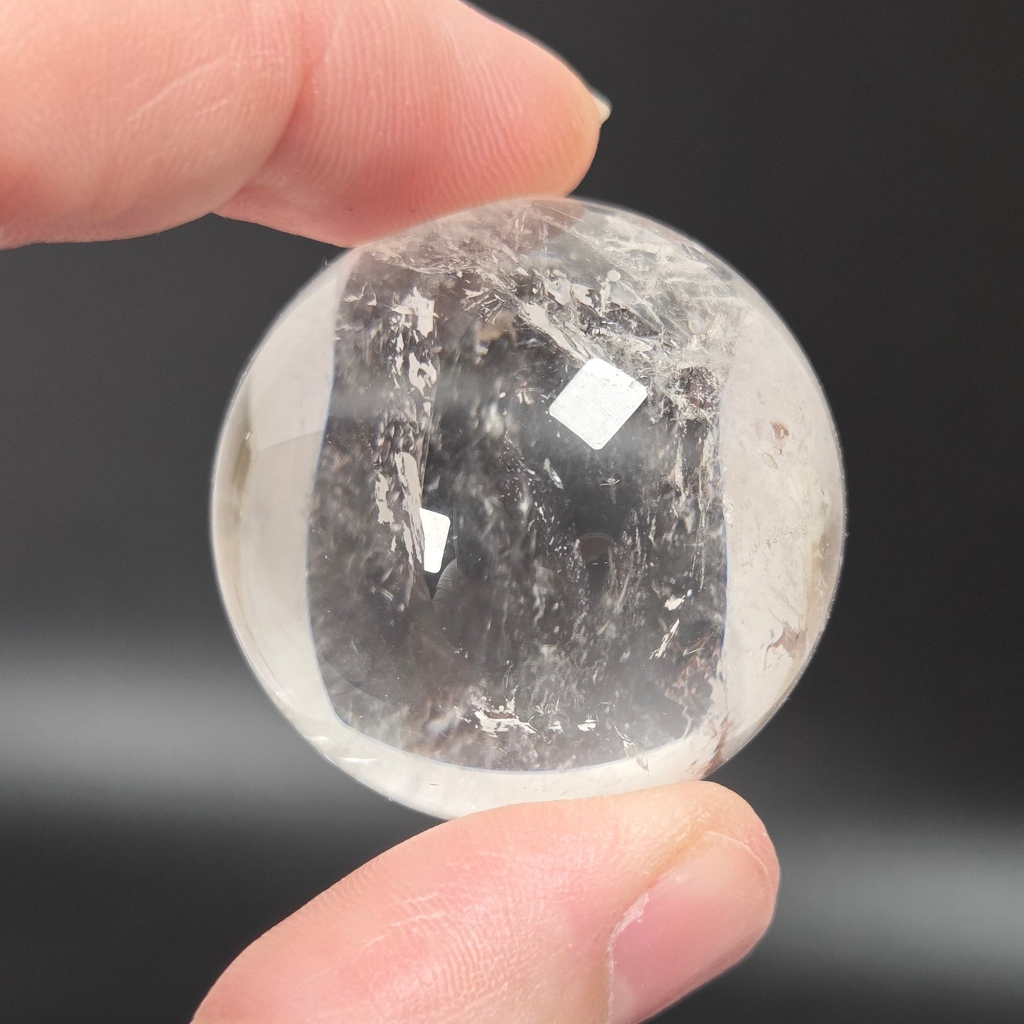 A hand holding a small, clear, faceted crystal sphere with internal inclusions against a dark gray background.