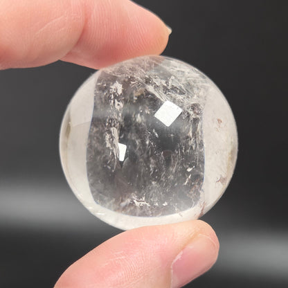 A hand holding a small, clear, faceted crystal sphere with internal inclusions against a dark gray background.