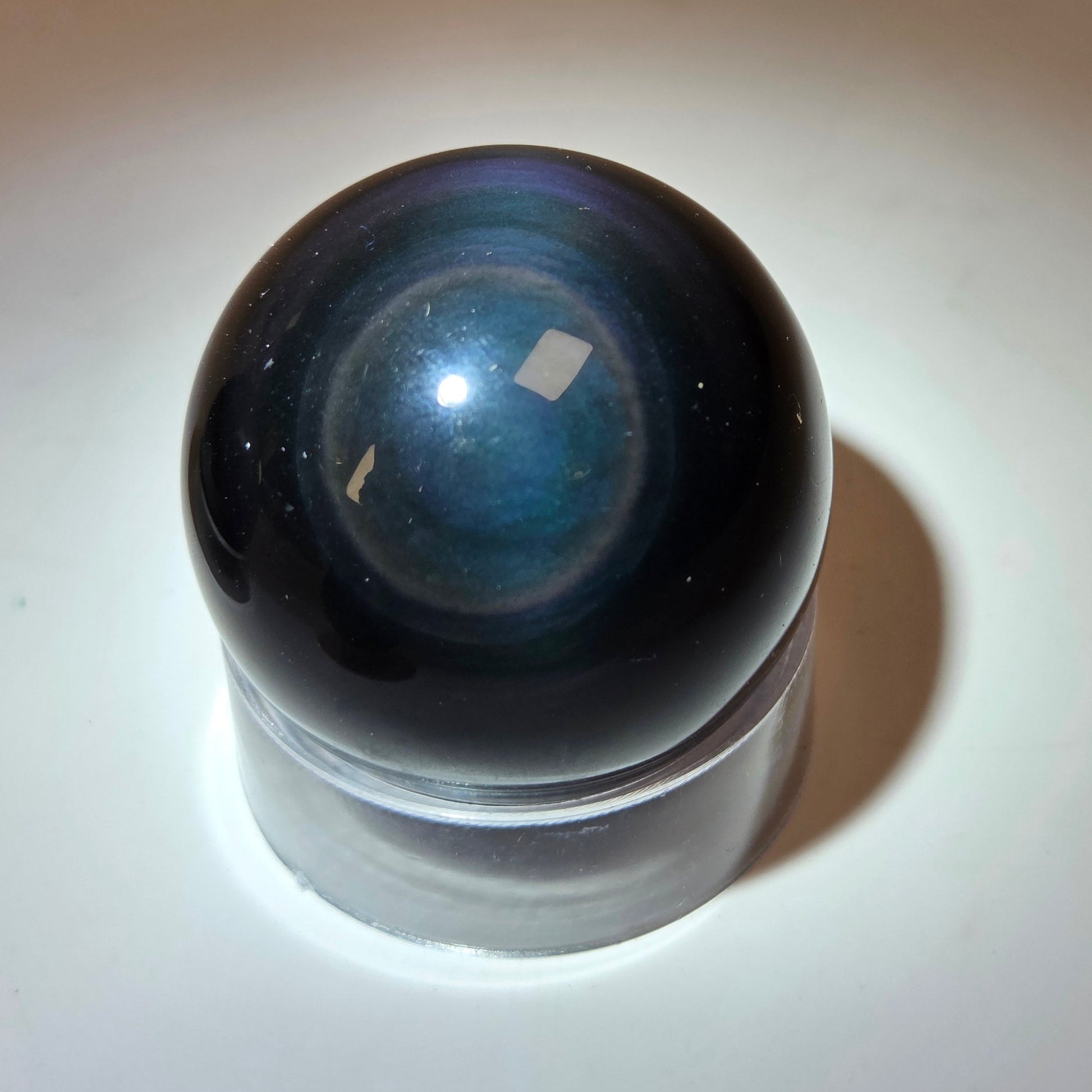 A polished dark blue and black glass sphere resting on a clear cylindrical base, photographed on a white surface with a warm beige background.