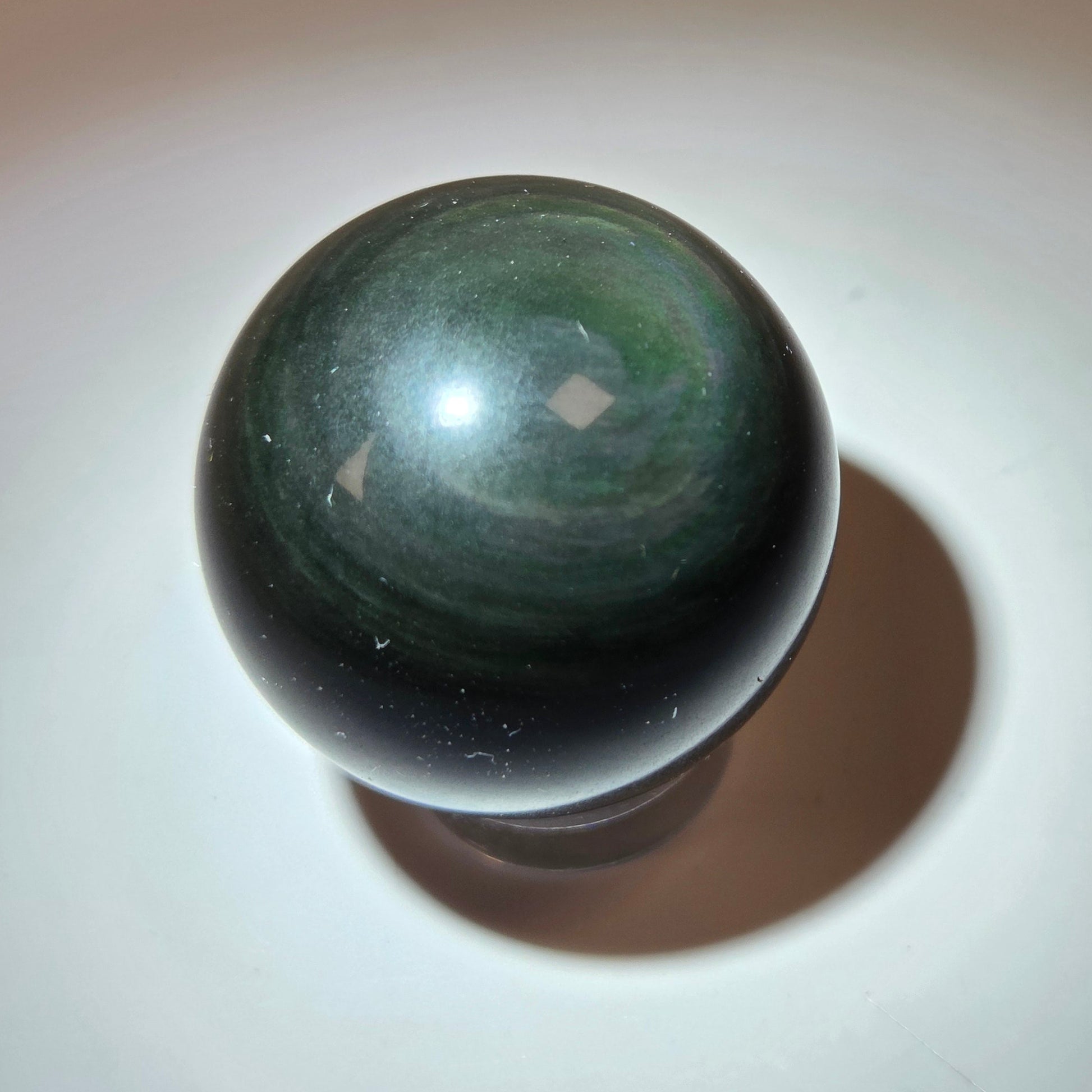 A polished dark green and black stone sphere, likely a tiger's eye or jasper bead, resting on a white surface with a warm spotlight and soft shadow.