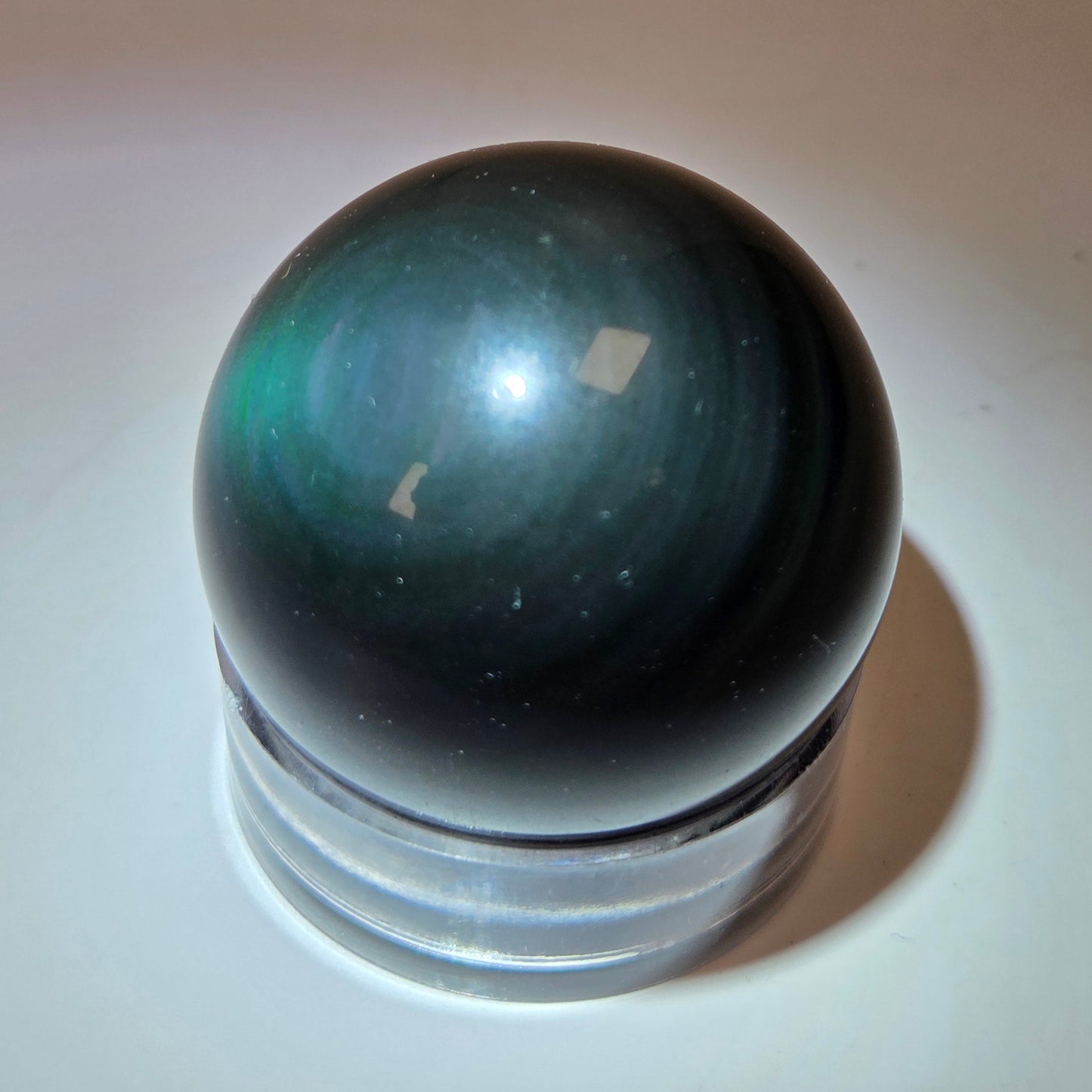 A polished dark green iridescent crystal sphere resting on a clear acrylic display stand, set against a plain beige background with a spotlight effect.