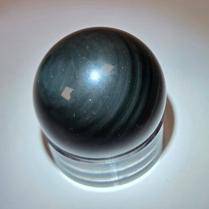 A polished black stone sphere with subtle greenish-gray banding, resting on a clear acrylic display base against a plain beige background.