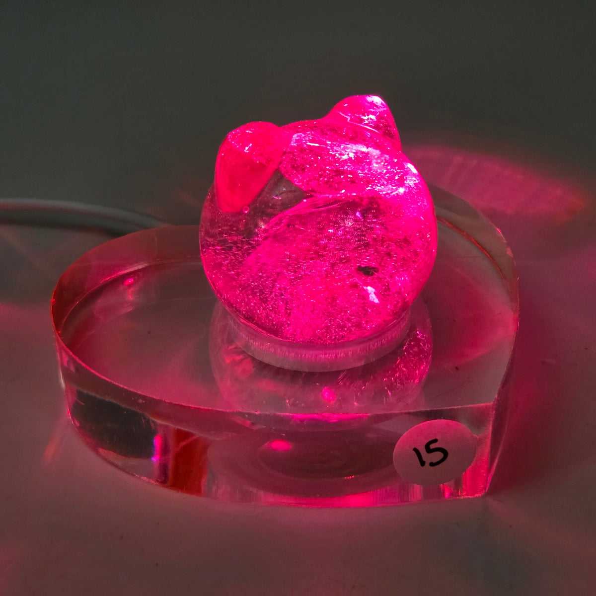 A small pink cat-shaped acrylic figurine with a heart-shaped head, displayed on a clear heart-shaped acrylic base. The item is photographed against a dark background, casting a strong pink reflection on the surface below.