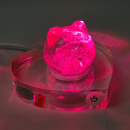 A small pink cat-shaped acrylic figurine with a heart-shaped head, displayed on a clear heart-shaped acrylic base. The item is photographed against a dark background, casting a strong pink reflection on the surface below.