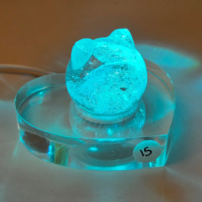 A glowing turquoise cat-shaped decorative object resting on a clear acrylic heart-shaped base, photographed on a warm tan surface with light reflections around it.