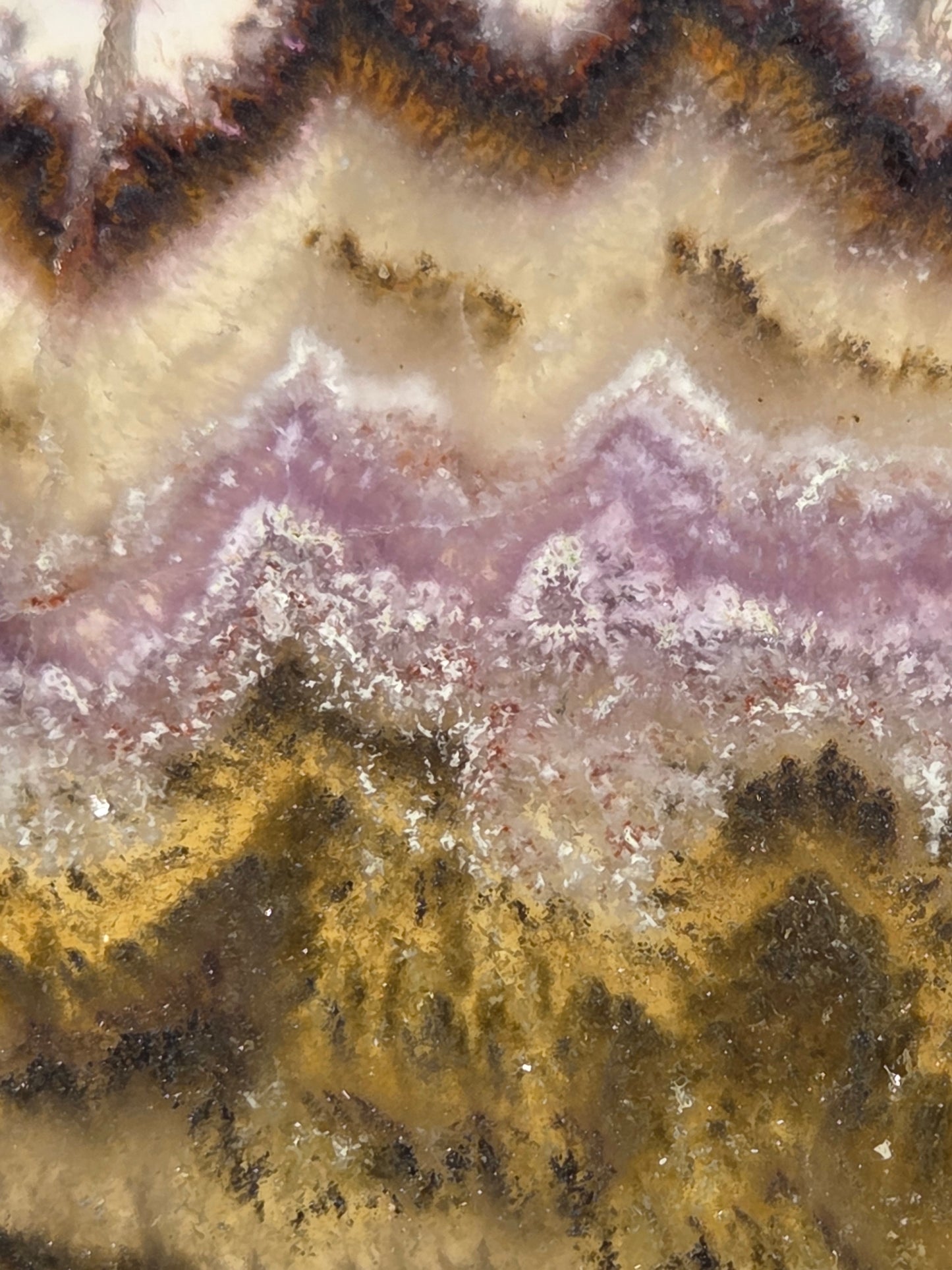 A close-up of a mineral or stone surface with layered bands of cream, lavender-purple, metallic gold, and dark brown, resembling agate or a polished geode cross-section.