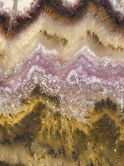 A close-up of a mineral or stone surface with layered bands of cream, lavender-purple, metallic gold, and dark brown, resembling agate or a polished geode cross-section.