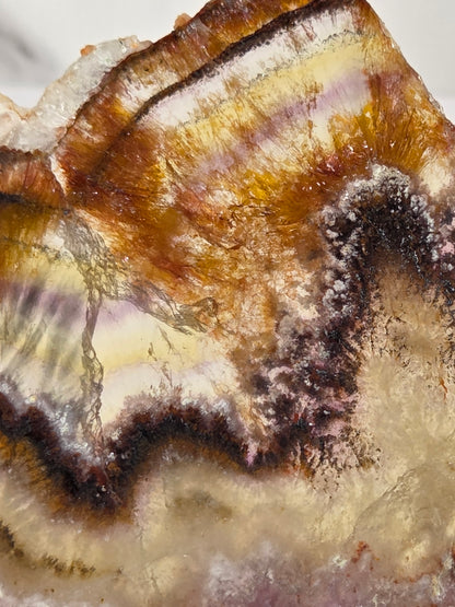 A close-up of a polished agate or quartz crystal specimen with layered bands of amber brown, cream, pale yellow, and dark purple-brown crystalline edges.